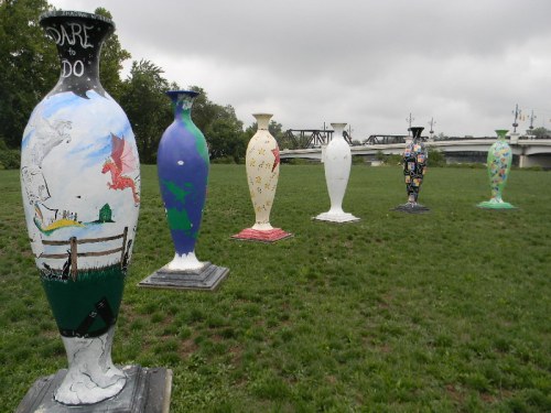 Colorful, artistic vases displayed on grass, with a cloudy sky and a bridge in the background.