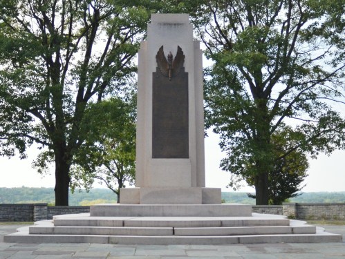 A tall stone monument with a bronze plaque, surrounded by trees and overlooking a scenic landscape.