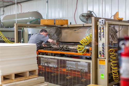 A worker operates machinery in a factory, surrounded by equipment and fans, focusing on a task.
