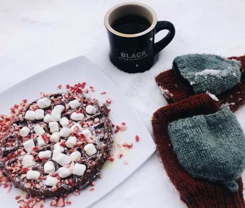 A cozy winter scene featuring a plate of dessert topped with marshmallows and crushed candy canes, alongside a coffee mug and mittens.