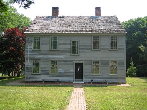 A two-story gray wooden house with a gabled roof, surrounded by grass and trees, featuring multiple windows and a front path.