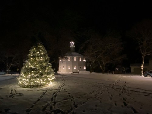 A snow-covered scene at night featuring a lit Christmas tree and a charming building in the background.