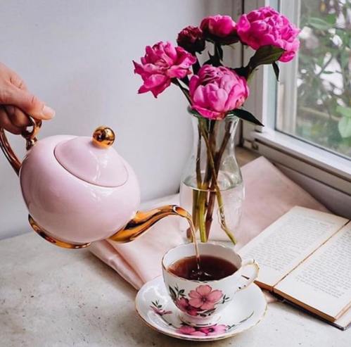 A hand pours tea from a pink teapot into a floral teacup, beside a vase of pink peonies and an open book.
