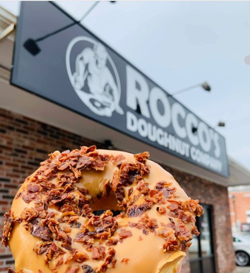 A close-up of a donut topped with caramel and crispy bacon, in front of Rocco's Doughnut Company sign.