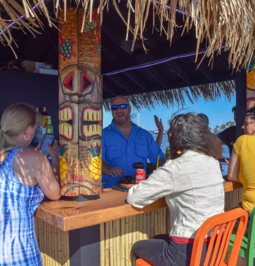 A tiki bar scene with a bartender smiling and chatting with customers under a thatched roof. Bright colors and tropical decor.