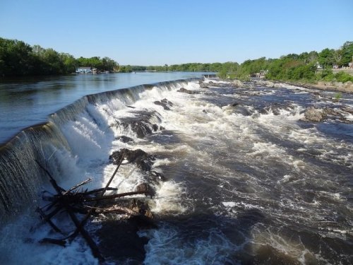 A scenic view of a river with a waterfall, surrounded by greenery and clear blue skies.