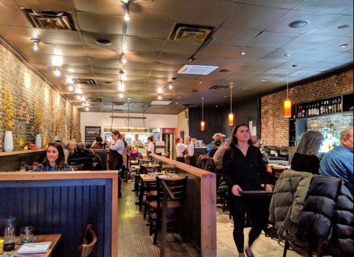 A bustling restaurant interior with diners, a server carrying a tray, and warm lighting highlighting brick walls.
