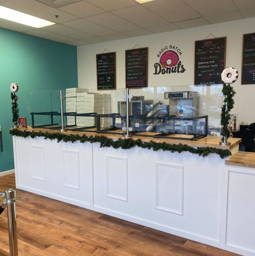 A bright donut shop counter with a glass barrier, decorated with garland, and a menu on the wall.