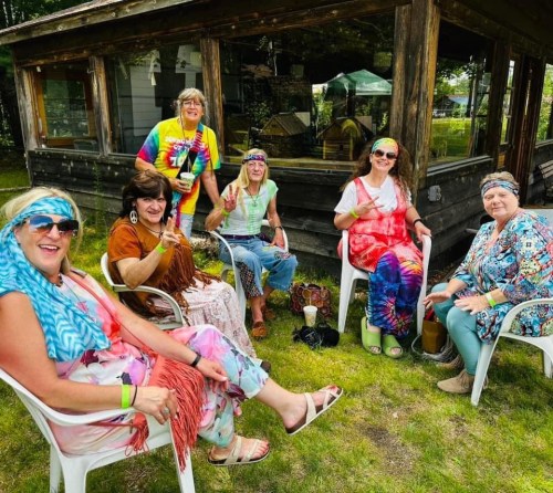 A group of six women in colorful outfits and headbands sit together outside, smiling and making peace signs.