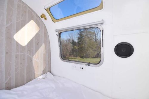 Cozy interior of a camper with a window showing a view of trees and blue sky, featuring a white bed and wall decor.