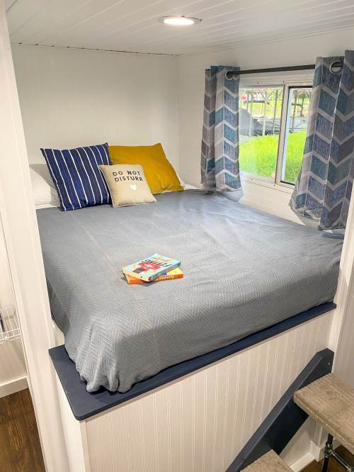 Cozy bedroom with a gray bedspread, decorative pillows, a book on the bed, and large windows with curtains.