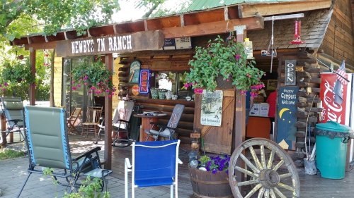 A rustic ranch storefront with hanging flowers, open sign, and outdoor seating, surrounded by greenery.