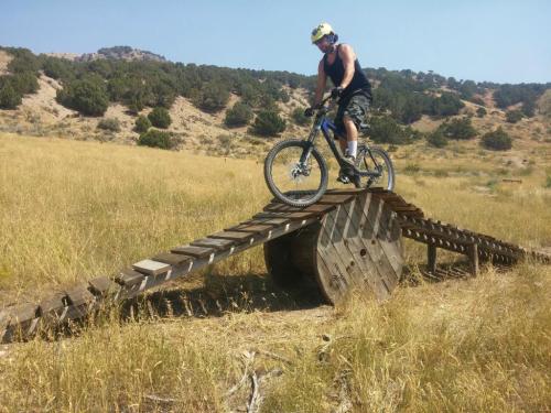 A cyclist balances on a wooden ramp over a large spool in a grassy field, surrounded by hills and trees.