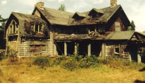 Abandoned, dilapidated house with overgrown vegetation and weathered wooden exterior under a cloudy sky.