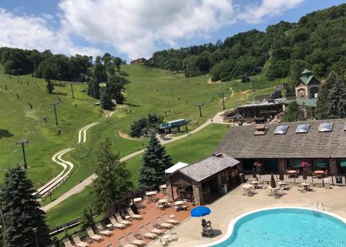 A scenic view of a ski resort with green hills, ski lifts, and a pool area surrounded by trees and blue skies.