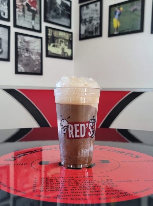 A frothy root beer float in a clear cup sits on a red vinyl table, with black-and-white photos on the wall behind it.