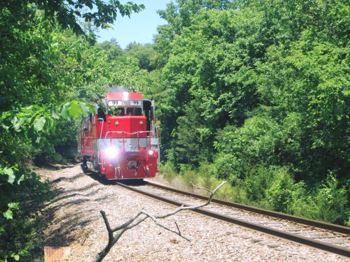 A red train approaches on a track surrounded by lush green trees and foliage.