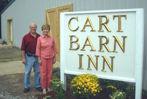 A man and woman stand beside a large sign that reads "CART BARN INN," with flowers in front.