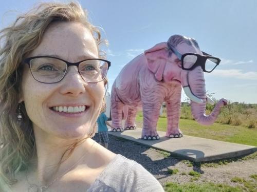 A smiling person with glasses poses next to a large, pink elephant statue wearing glasses in a sunny outdoor setting.