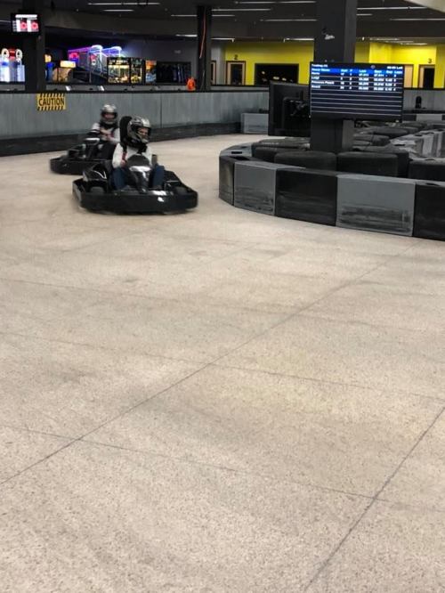 Two go-karts racing on a track inside an indoor facility, with a scoreboard visible in the background.
