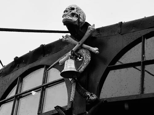 A skull and crossbones decoration with a bell and snake, mounted on a dark building with windows.