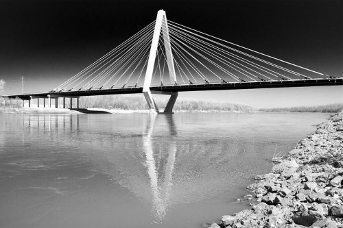 A modern cable-stayed bridge spans a river, reflected in the water, with rocky banks visible in the foreground.