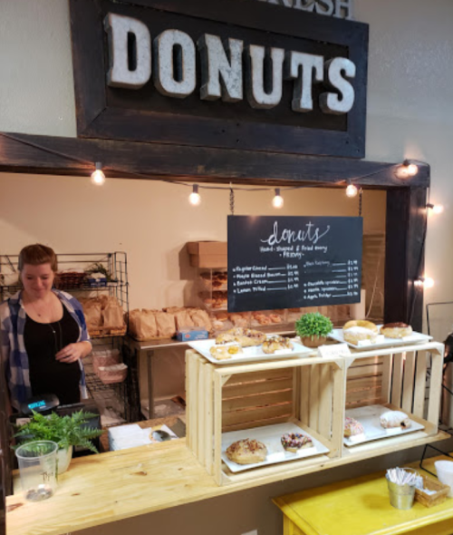 A cozy donut shop with a menu board, fresh donuts on display, and a smiling employee behind the counter.