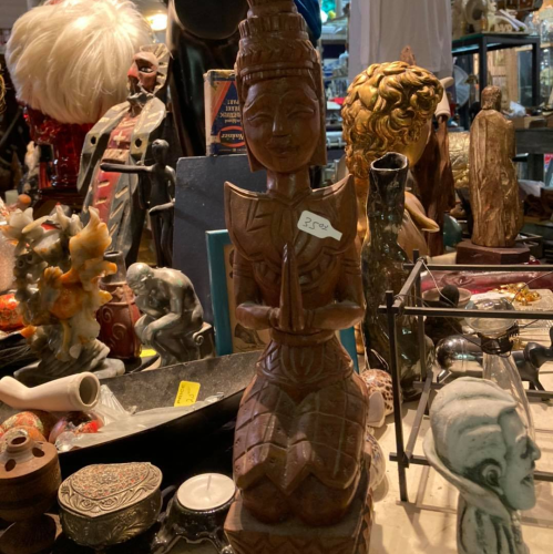 A wooden statue of a figure with intricate carvings, surrounded by various antiques and collectibles on a cluttered table.