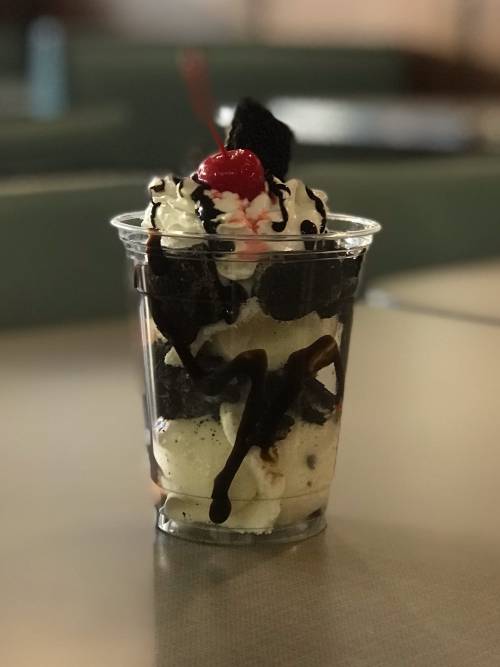 A clear cup filled with layers of ice cream, whipped cream, chocolate sauce, crushed cookies, and a cherry on top.