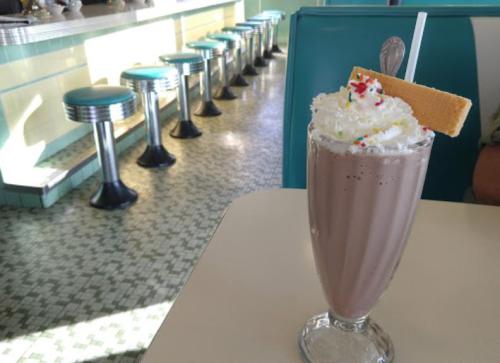 A chocolate milkshake topped with whipped cream and sprinkles, served in a glass with a wafer, in a retro diner setting.