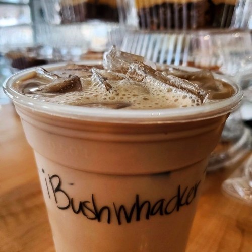 A close-up of a iced coffee drink in a clear cup, labeled "Bushwhacker," with ice cubes on top.
