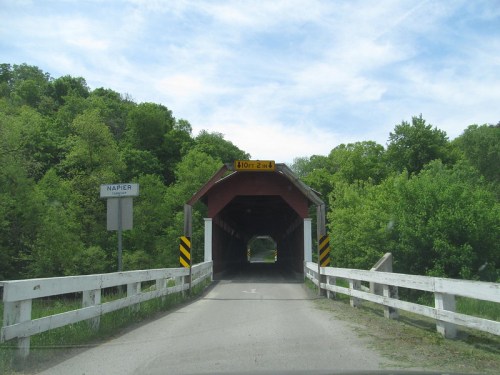 A covered bridge with a red exterior, surrounded by green trees, leading to a sign indicating "Napier."