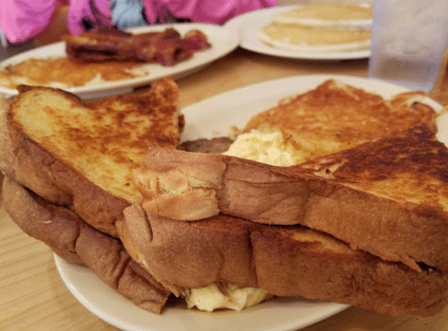A plate of French toast stacked with scrambled eggs, alongside pancakes and bacon in the background.