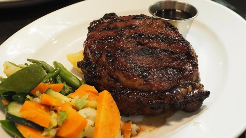 A grilled steak served with a side of mixed vegetables and a small cup of sauce on a white plate.