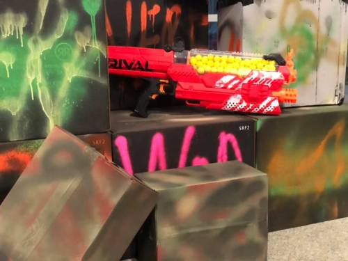 A brightly colored toy blaster rests against spray-painted boxes in a playful, urban setting.