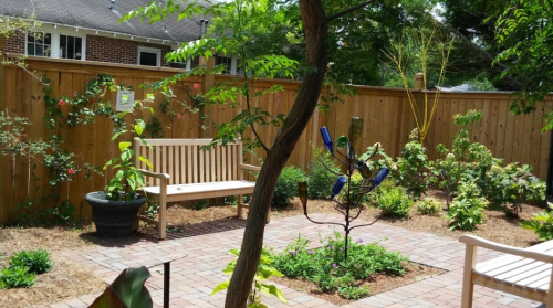 A serene garden with a wooden bench, colorful plants, and a decorative sculpture surrounded by a wooden fence.