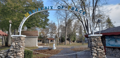 Archway sign reading "Mediatrix of Peace" in a park-like setting with trees and stone structures.