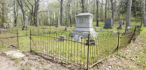 A historic cemetery with a fenced grave surrounded by trees and headstones, featuring a prominent stone monument.