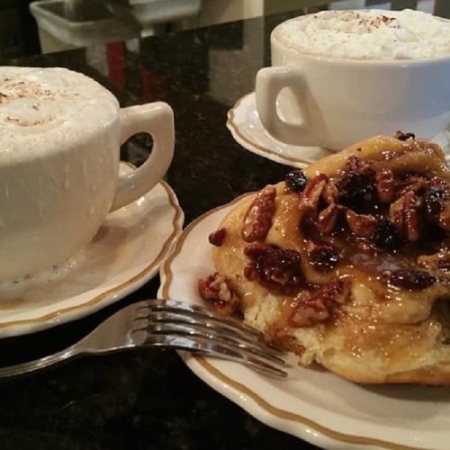 Two cups of frothy coffee and a cinnamon roll topped with nuts, served on white plates with a fork.
