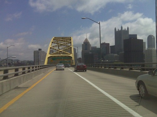 A yellow bridge spans a highway with cars, leading to a city skyline under a partly cloudy sky.