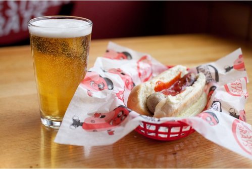 A glass of beer next to a hot dog in a red basket lined with patterned paper on a wooden table.