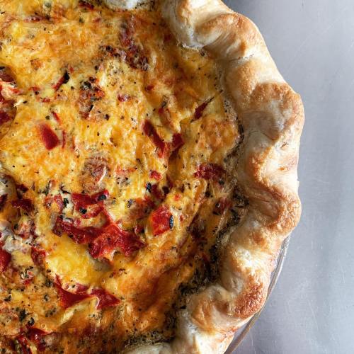 A freshly baked quiche with a golden crust, filled with cheese and red peppers, on a silver surface.