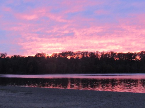 A serene lake at sunset, reflecting vibrant pink and purple hues in the sky, with trees silhouetted along the horizon.