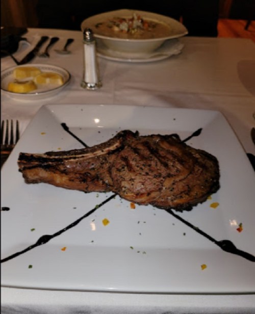 A plated ribeye steak with grill marks, garnished with sauce and herbs, served on a white square dish.