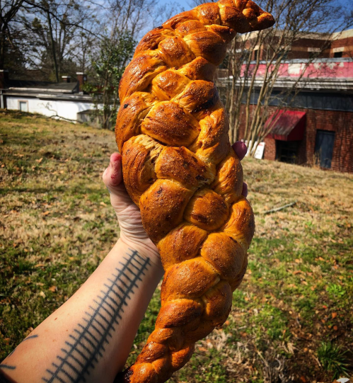 A hand holds a large, braided loaf of bread against a grassy background.