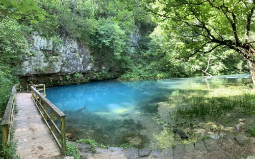 A serene blue pond surrounded by lush greenery and rocky cliffs, with a wooden walkway leading to the water's edge.