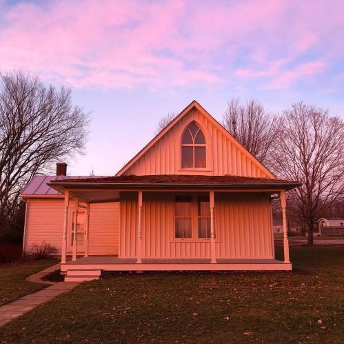 A charming white house with a peaked roof and large front porch, set against a pink and purple sunset sky.