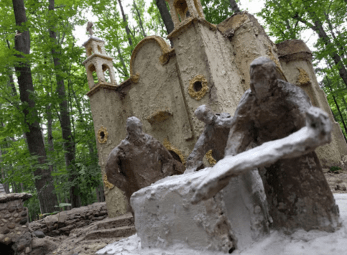 Sculptures of three figures working on a stone structure, set in a forest with greenery and a partially visible building.