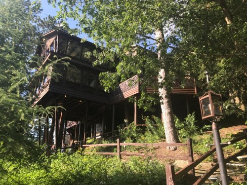 A large wooden house surrounded by trees, featuring multiple levels and a staircase leading up to it.