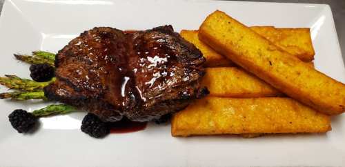 A plated meal featuring a grilled steak, polenta fries, asparagus, and blackberries drizzled with sauce.